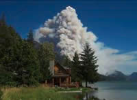 Picture of a dense white smoke column rising behind a cabin on the shoreline of a lake.