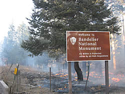 fire burning under the trees behind a Bandelier National Monument sign