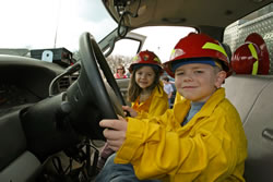 A young boy and girl wearing hard hats and fire shirts sitting in a truck.
