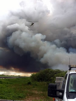 View along a firefighter's truck of a helicopter hovering with a smoke column in the background.